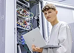 Fachinformatiker mit Laptop in der linken Hand steht vor einem geöffnetem Serverschrank und Blickt in die Kamera